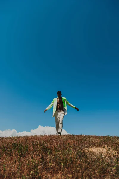 African american man in white trousers and blazer walking under blue sky in field - foto de stock