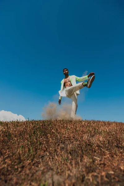 Афроамериканський чоловік у стильному одязі позує з піднятою ногою на лузі під блакитним небом — стокове фото
