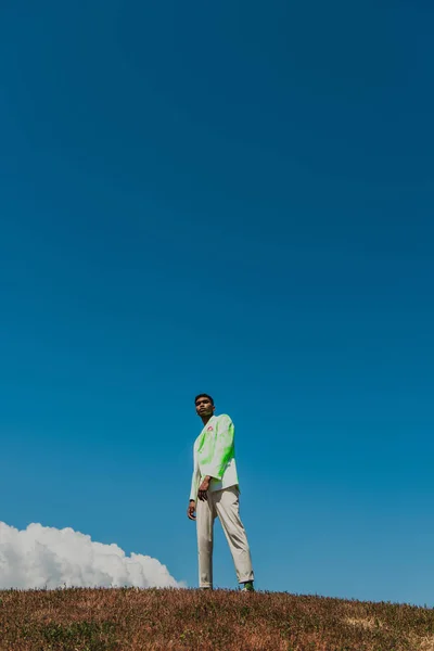 African american man in stylish blazer and trousers standing in meadow under blue sky — Stockfoto