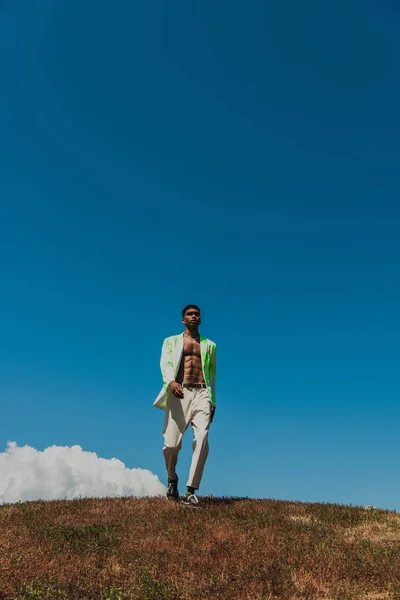 Full length of african american man in trousers and blazer walking in field under blue sky — Photo de stock