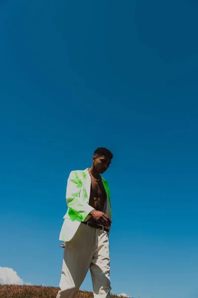 Young african american man in fashionable blazer walking under clear sky in summer — стоковое фото