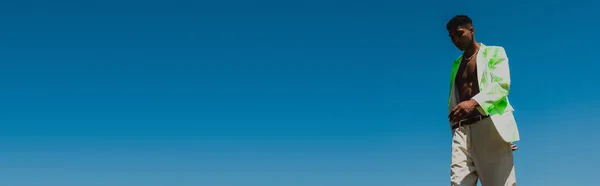 Young african american man in trendy white and green blazer against cloudless sky, banner - foto de stock