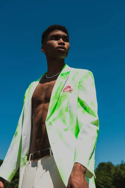 Low angle view of african american man in white and green blazer standing under blue sky and looking away - foto de stock