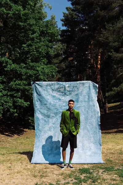 Full length of african american man in velvet blazer and shorts standing with hands in pockets near blue cloth in park — Stock Photo