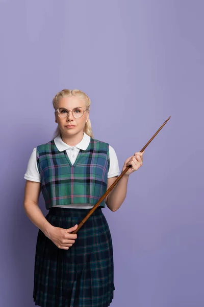 Blonde student in uniform and eyeglasses holding pointer isolated on purple - foto de stock