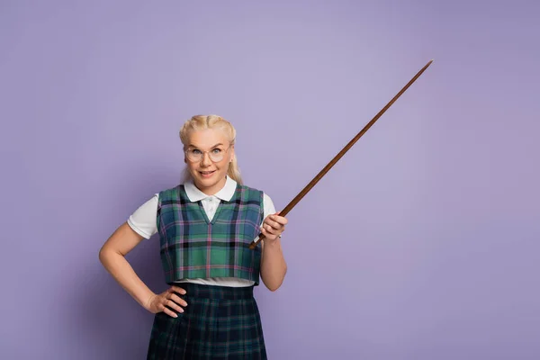 Smiling blonde woman in vest holding pointer and hand on hip on purple background - foto de stock