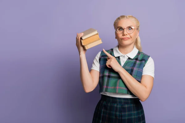 Confused student in eyeglasses pointing at books on purple background — Photo de stock