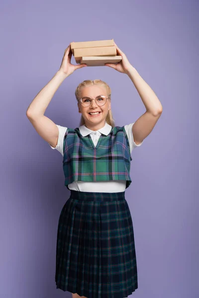 Cheerful blonde student in uniform holding books above head isolated on purple — Stock Photo