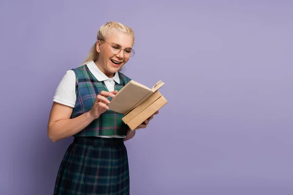 Positive student in eyeglasses reading book isolated on purple — Stock Photo