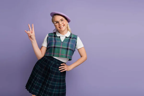 Smiling student in eyeglasses and beret showing peace gesture isolated on purple — Stockfoto