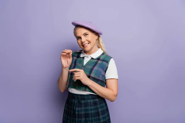 Cheerful student in uniform and beret holding eyeglasses on purple background — Foto stock