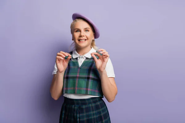 Positive blonde student in beret holding eyeglasses on purple background — Photo de stock