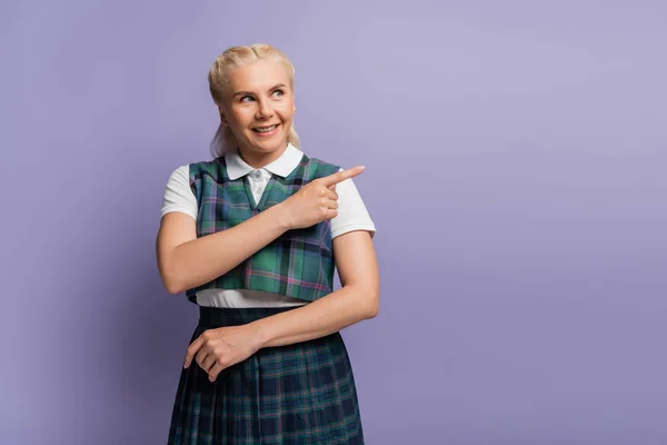 Smiling student in checkered uniform pointing with finger isolated on purple — стоковое фото