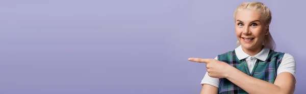 Happy blonde student pointing with finger isolated on purple with copy space, banner — Stockfoto