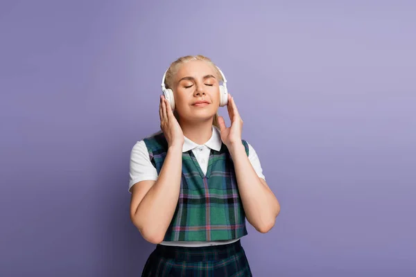 Student in checkered uniform listening music in headphones isolated on purple — стоковое фото