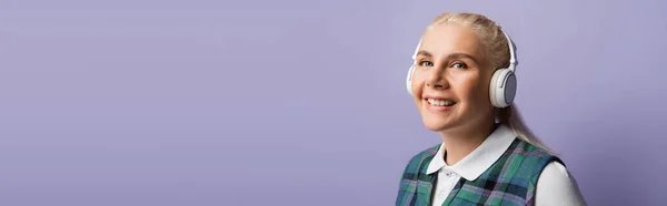 Positive blonde student in headphones looking at camera isolated on purple, banner — Foto stock