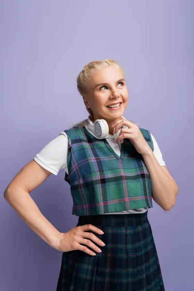 Positive student in uniform holding headphones isolated on purple — стоковое фото