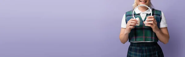 Cropped view of cheerful student in uniform holding wireless headphones isolated on purple, banner — Photo de stock