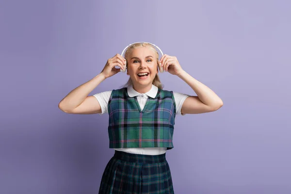 Positive student in uniform holding headphones near ears isolated on purple — стоковое фото