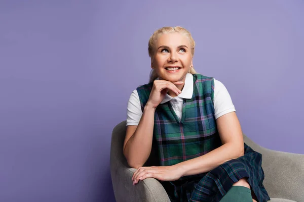Cheerful student in checkered uniform sitting on armchair isolated on purple — Stock Photo