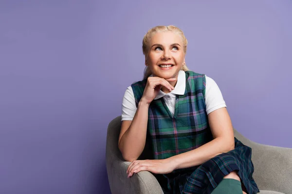 Cheerful student in checkered uniform sitting on armchair isolated on purple - foto de stock