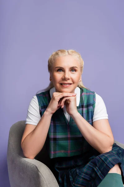 Positive student in vest and t-shirt looking at camera on armchair isolated on purple — Stockfoto