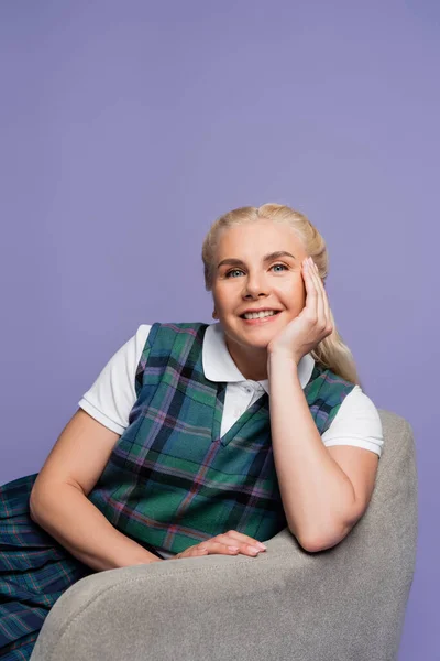 Happy blonde student in plaid vest sitting on armchair isolated on purple - foto de stock