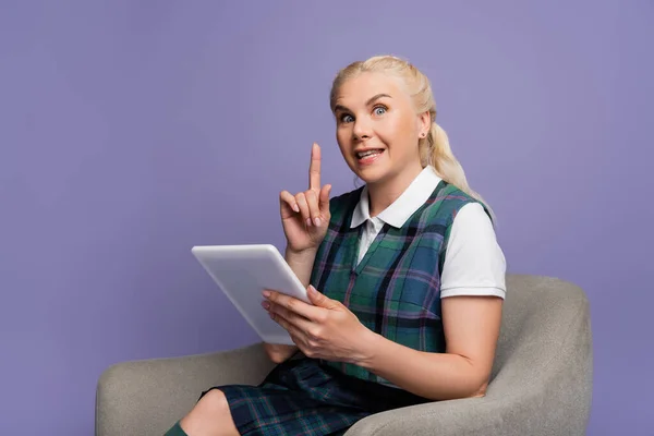 Excited student having idea while holding digital tablet on armchair isolated on purple - foto de stock