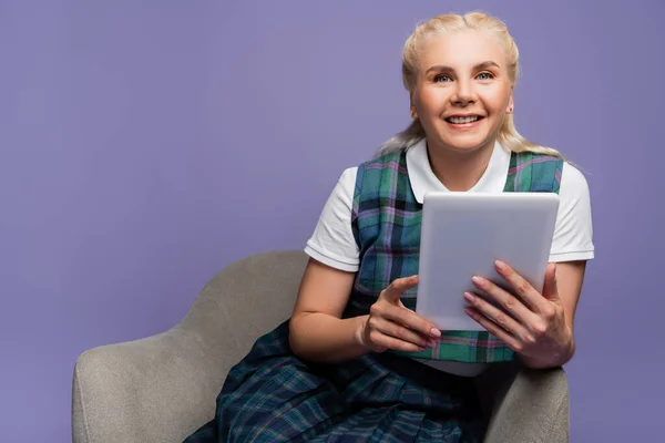 Positive student using digital tablet while sitting on armchair isolated on purple — Stockfoto