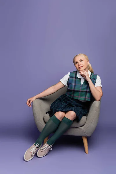 Dreamy student in plaid uniform sitting on armchair on purple background — Photo de stock