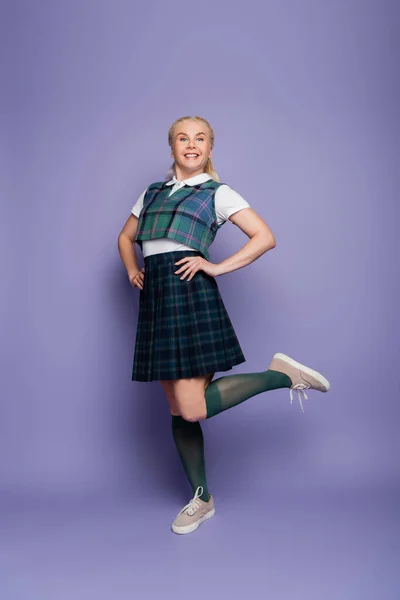 Positive student in checkered uniform holding hands on hips on purple background — Fotografia de Stock