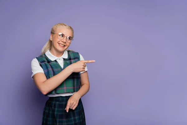 Smiling student in uniform and eyeglasses pointing with finger isolated on purple — Stockfoto