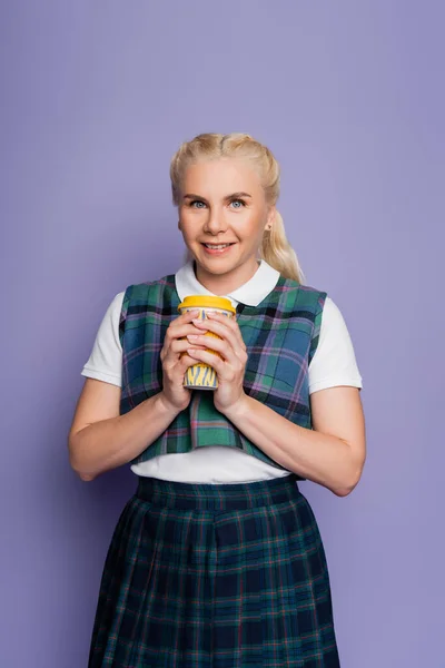 Positive student in plaid uniform holding coffee to go isolated on purple - foto de stock