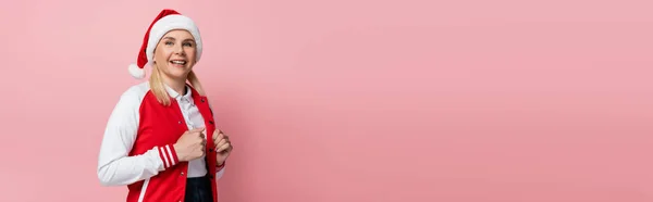 Cheerful woman in santa hat looking at camera isolated on pink, banner — стоковое фото