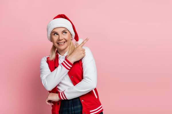 Cheerful woman in santa hat and baseball jacket pointing with finger isolated on pink — стоковое фото