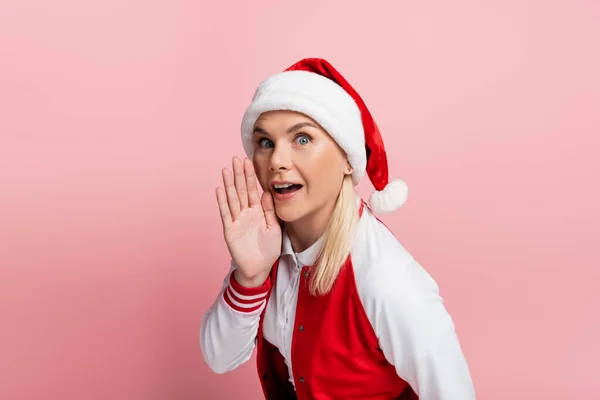 Excited woman in santa hat holding hand near cheek isolated on pink — Stock Photo