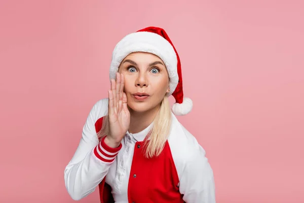 Astonished woman in santa hat holding hand near cheek isolated on pink — Foto stock