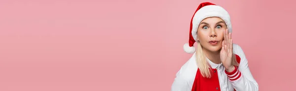 Amazed woman in santa hat holding hand near cheek isolated on pink, banner — Stock Photo