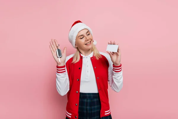 Cheerful woman in santa hat holding car key and empty driving license isolated on pink — стоковое фото