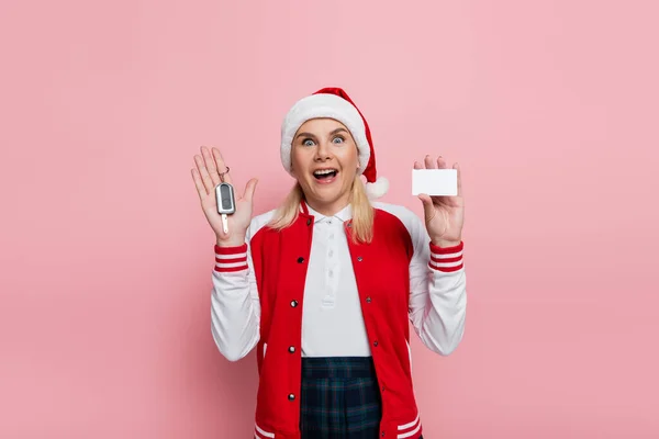 Excited woman in santa hat holding car key and empty driving license isolated on pink - foto de stock