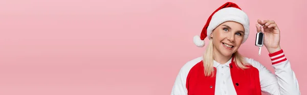 Positive woman in santa hat and baseball jacket holding car key isolated on pink, banner — Stockfoto