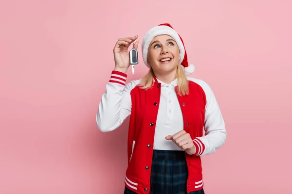 Positive woman in red santa hat holding car key and looking up isolated on pink - foto de stock