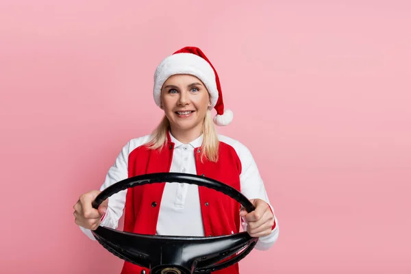 Smiling woman in santa hat holding steering wheel and looking at camera isolated on pink — Stock Photo