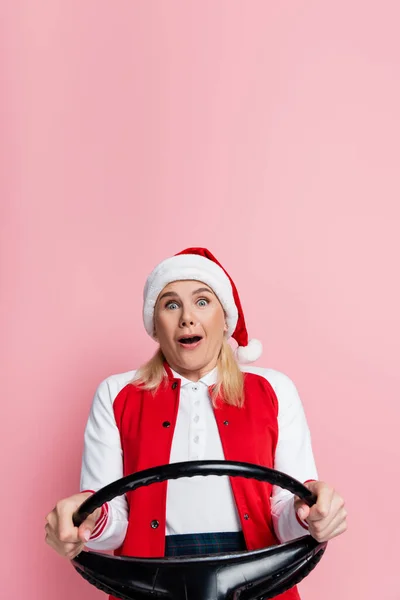 Scared woman in santa hat holding steering wheel isolated on pink - foto de stock