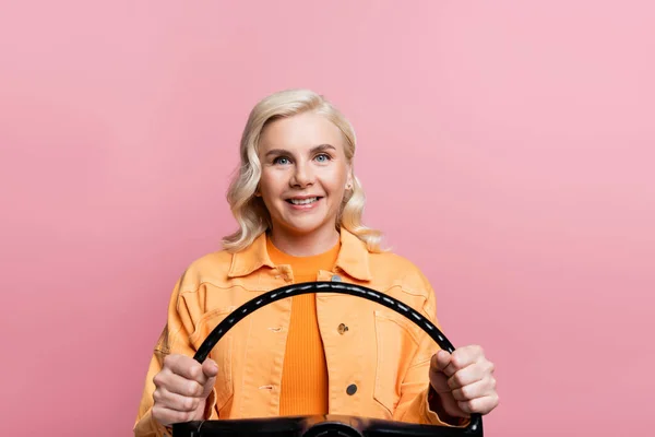 Happy blonde woman looking at camera while holding steering wheel isolated on pink — Stock Photo