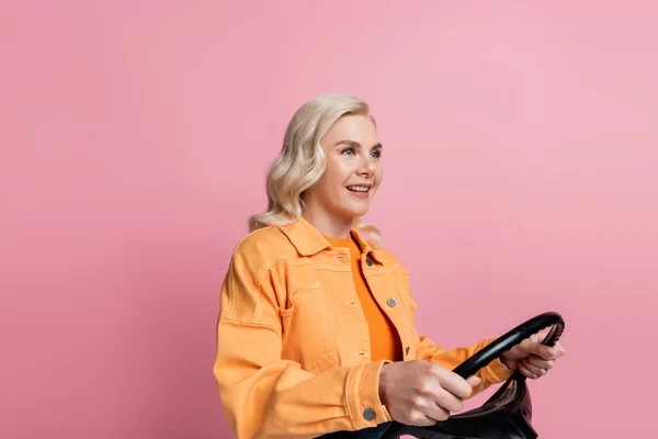 Blonde driver smiling and holding steering wheel isolated on pink — Stockfoto