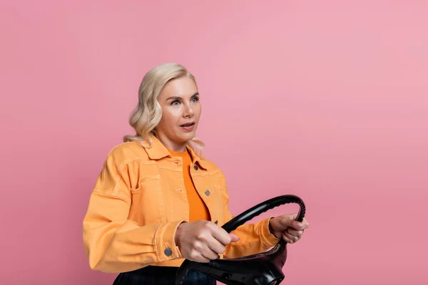 Blonde driver in orange jacket holding steering wheel isolated on pink — стоковое фото