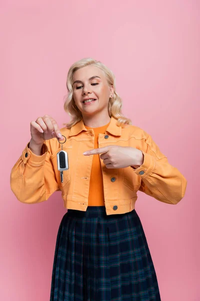 Smiling blonde woman pointing at car key isolated on pink — стоковое фото