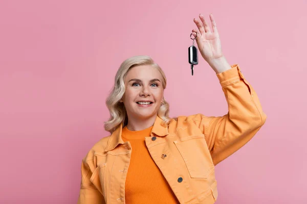 Positive driver in orange jacket holding car key isolated on pink — Stock Photo