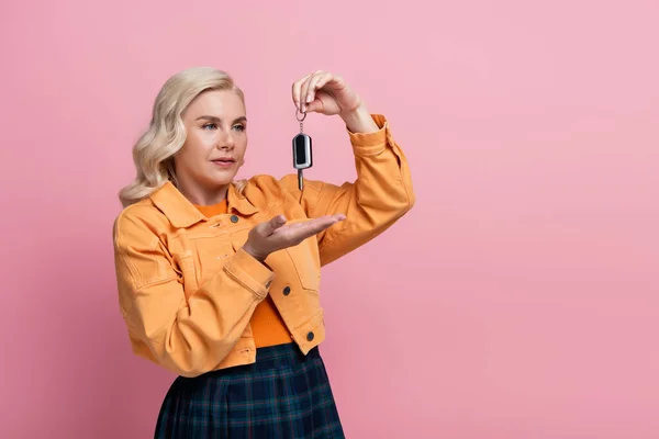 Блондинка в куртці вказує на ключ автомобіля ізольовано на рожевому — стокове фото